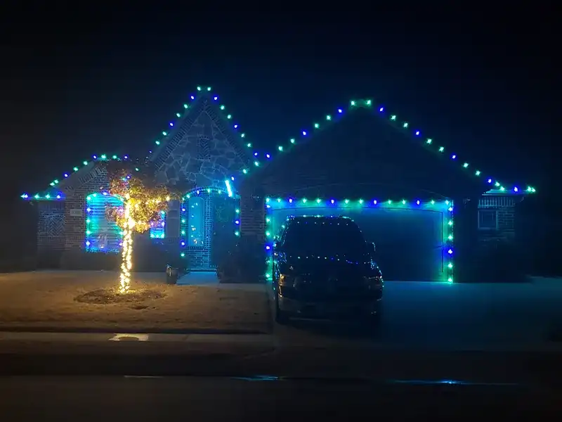 Home outlined in blue and green Christmas lights with a lit tree in the yard