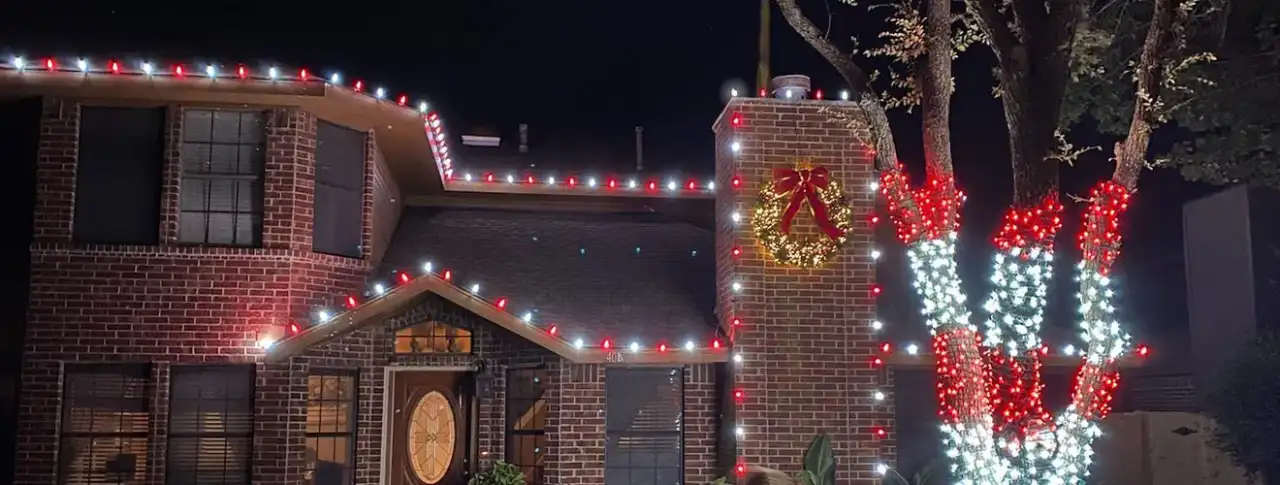 Brick home with a lit chimney wreath and red and white wrapped trees